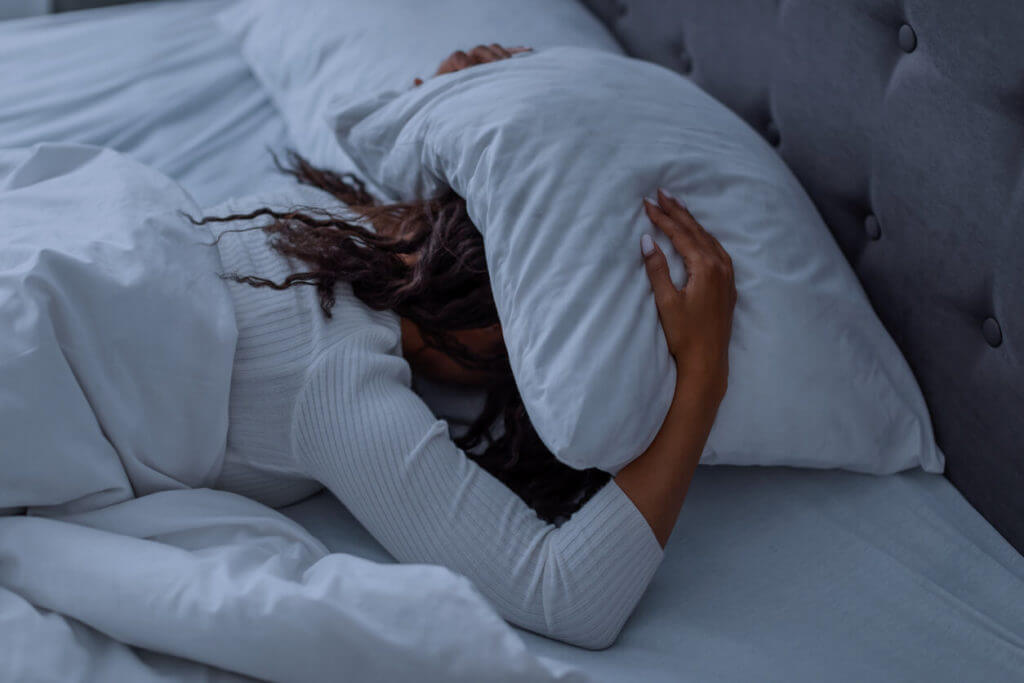 A stressed Black woman is lying in bed, covering her head with a pillow due to a sleep disorder.