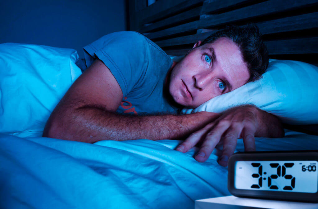 A restless, worried young man lies awake at night, suffering from insomnia and feeling depressed and sad due to stress and lack of sleep.